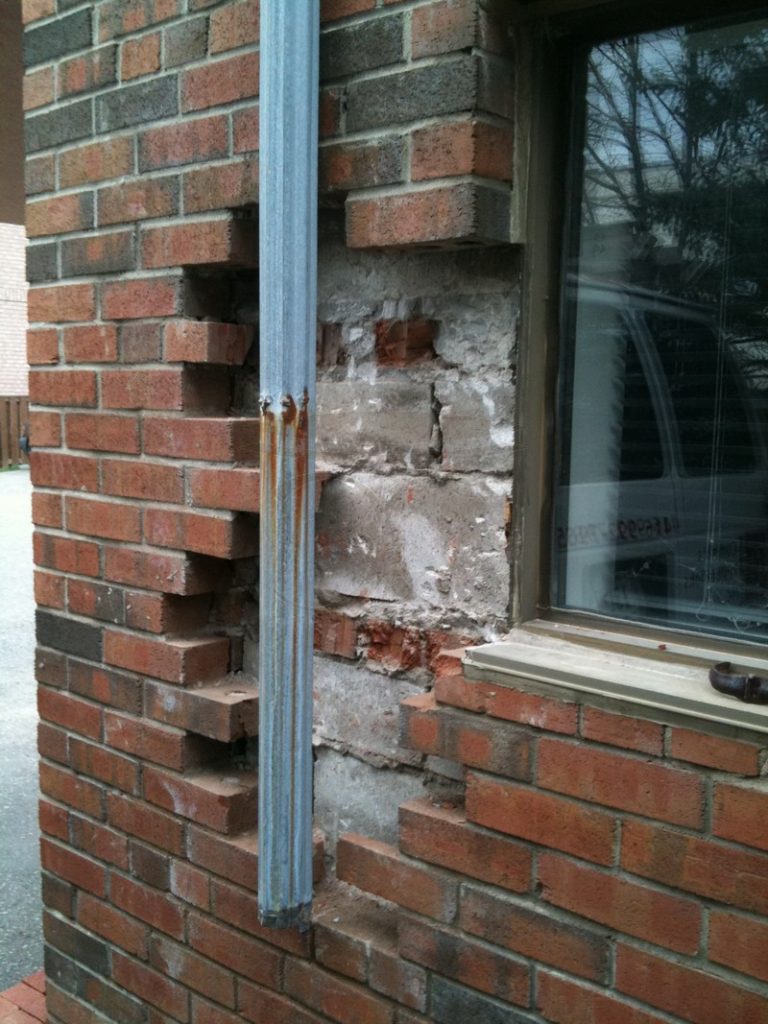 Brick wall beside a window with a steel lintel and missing bricks along the downspout