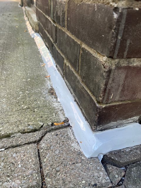 Base of a dark brick wall at a concrete walkway, with new membrane flashing along the joint