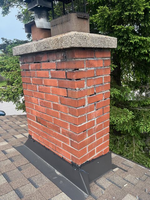 Brick chimney on a shingled roof with worn, open mortar joints below the concrete crown