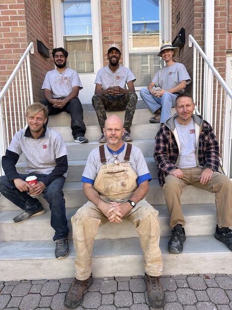 Five members of the Cummins Restorations crew in matching grey work shirts sitting together on freshly finished concrete steps in front of a brick townhouse