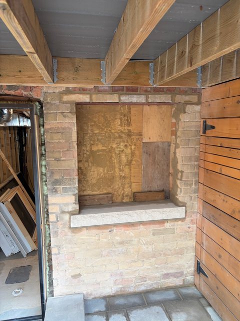 Buff brick wall under a deck with a boarded window opening and a new stone sill