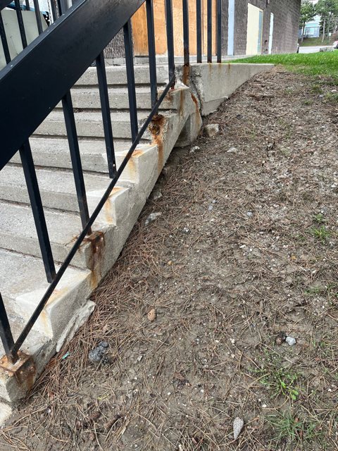 Crumbling concrete side wall along exterior steps with rust stains from the black metal railing posts